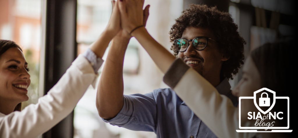 Three people are smiling and engaged in a celebratory high-five, with their hands raised together in a lively and joyful moment. The setting appears to be an office or collaborative space, indicating teamwork and positive interaction.