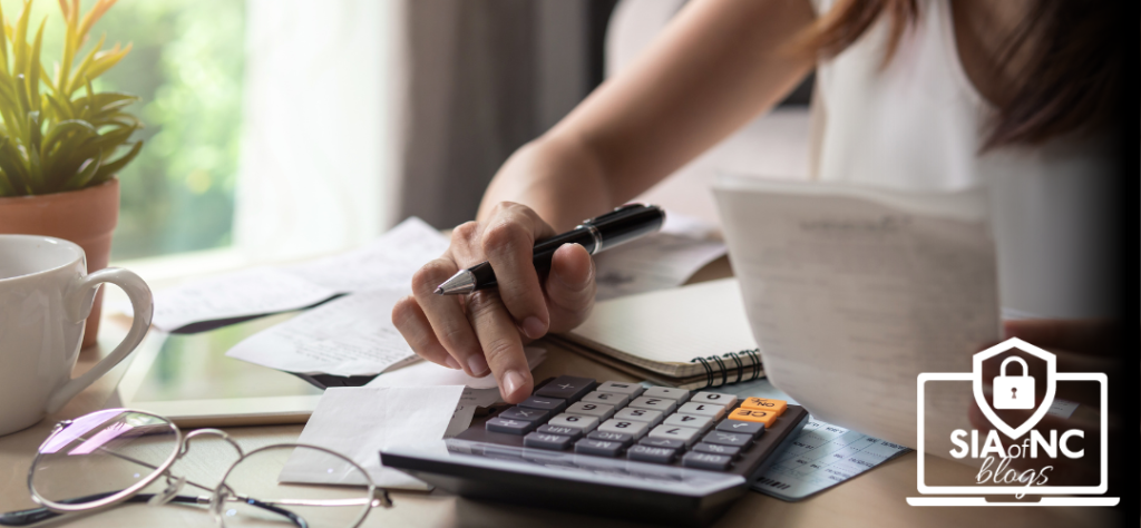 A person is using a calculator while reviewing documents and notes at a desk, with a cup of coffee and eyeglasses nearby. The image includes the SIA of NC Blogs logo in the bottom-right corner.